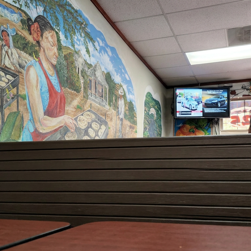 Restaurant dining area with wooden booth seating, colorful mural of food preparation, fruit decorations, and a wall-mounted television.